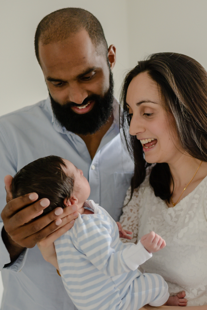 Photo of parents holding newborn baby boy, both smile down at baby who is sleeping